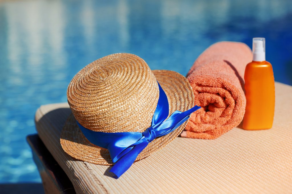 Orange cotton towel and sunscreen body lotion and hat in a orange tube on the background pool. Means for skin care, concept of relaxation and sun protection.